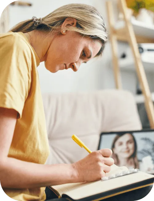 A person wearing a yellow shirt writing in a notebook with a yellow pen. In front of them, a tablet or laptop screen displays a video call with another person who is speaking.